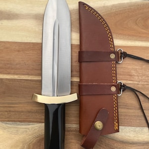 Pu&ograve; includere: Un pugnale d'argento con manico nero e guardia in ottone. Il pugnale &egrave; in un fodero di cuoio marrone con un bottone in ottone e cuciture.