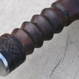 Pu&ograve; includere: Primo piano di un manico in legno marrone scuro con un design a spirale. Il manico ha un'estremit&agrave; in metallo argentato con una punta e una fascia decorativa. La venatura del legno &egrave; visibile e il manico sembra far parte di uno strumento.