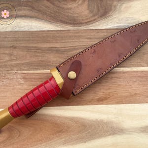 Peut inclure: Un couteau &agrave; manche rouge avec une base dor&eacute;e et un &eacute;tui en cuir marron. L'&eacute;tui est fix&eacute; avec une lani&egrave;re en cuir et un bouton dor&eacute;. Le couteau repose sur une surface en bois.