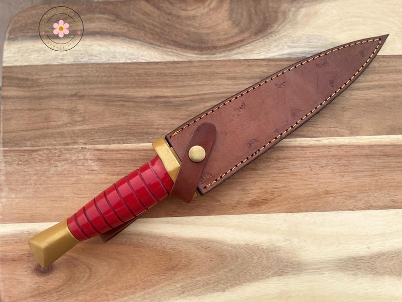 Peut inclure: Un couteau &agrave; manche rouge avec une base dor&eacute;e et un &eacute;tui en cuir marron. L'&eacute;tui est fix&eacute; avec une lani&egrave;re en cuir et un bouton dor&eacute;. Le couteau repose sur une surface en bois.
