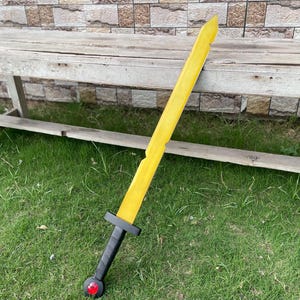 May include: A yellow toy sword with a black handle and a red gem at the base. The sword is lying on green grass next to a wooden bench. The sword's blade has a slightly jagged edge.