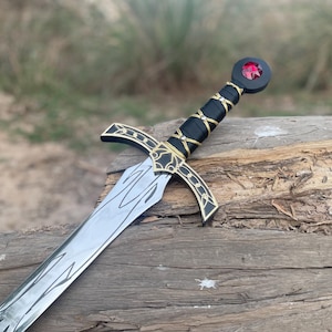 Op de afbeelding: Een decoratief zwaard met een zilveren lemmet en een zwarte handgreep omwikkeld met zwart leer. De handgreep heeft gouden accenten en een rode edelsteen op de pommel. Het zwaard ligt op een verweerd houten oppervlak.