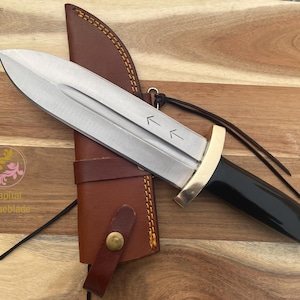 Pu&ograve; includere: Un coltello argentato con manico nero e guardia dorata. Il coltello ha un fodero in pelle con una cinghia marrone. Il coltello ha due frecce che puntano verso l'alto sulla lama.