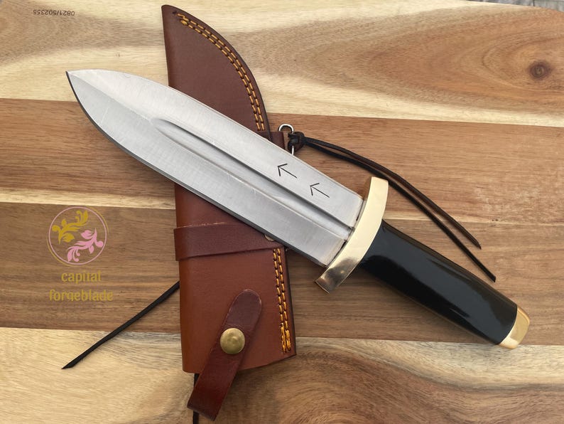 Pu&ograve; includere: Un coltello argentato con manico nero e guardia dorata. Il coltello ha un fodero in pelle con una cinghia marrone. Il coltello ha due frecce che puntano verso l'alto sulla lama.