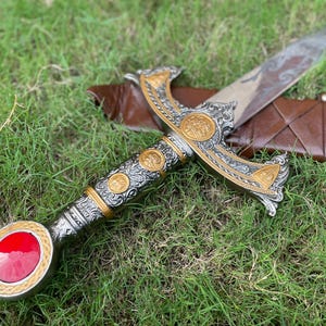 May include: An ornamental sword with a silver and gold handle, featuring a red gem at the pommel. The blade is etched with a design, and the sword rests on a brown leather sheath, set against a backdrop of green grass.