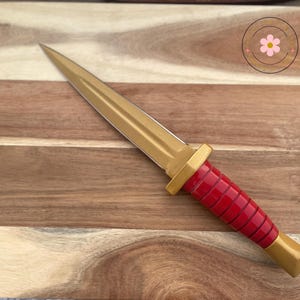 Peut inclure: Un poignard d&eacute;coratif avec une lame et une poign&eacute;e dor&eacute;es, et une poign&eacute;e rouge. Le poignard est pos&eacute; sur une planche &agrave; d&eacute;couper en bois.