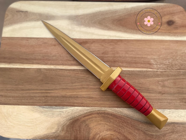 Peut inclure: Un poignard d&eacute;coratif avec une lame et une poign&eacute;e dor&eacute;es, et une poign&eacute;e rouge. Le poignard est pos&eacute; sur une planche &agrave; d&eacute;couper en bois.