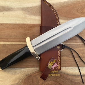 Pu&ograve; includere: Un coltello con manico nero, lama argentata e guardia dorata. Il coltello &egrave; in un fodero di cuoio marrone con un motivo floreale decorativo e il testo "capital forgeblade".