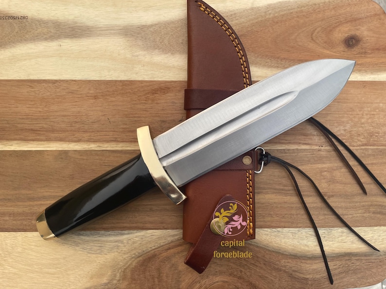 Pu&ograve; includere: Un coltello con manico nero, lama argentata e guardia dorata. Il coltello &egrave; in un fodero di cuoio marrone con un motivo floreale decorativo e il testo "capital forgeblade".