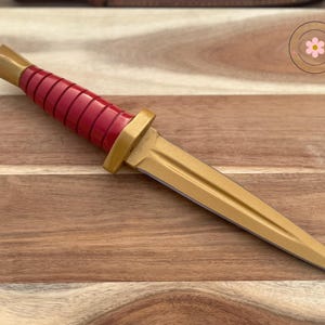 Peut inclure: Un poignard d&eacute;coratif avec une lame et des accents dor&eacute;s. Le manche est rouge avec un design nervur&eacute;. Le poignard mesure environ 25 cm de long et repose sur une surface en bois.