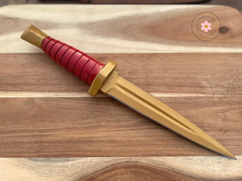 Peut inclure: Un poignard d&eacute;coratif avec une lame et des accents dor&eacute;s. Le manche est rouge avec un design nervur&eacute;. Le poignard mesure environ 25 cm de long et repose sur une surface en bois.