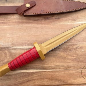 Peut inclure: Un poignard dor&eacute; avec un manche rouge et un &eacute;tui en cuir marron. Le poignard a une lame pointue et un manche d&eacute;coratif. L'&eacute;tui est fix&eacute; avec une sangle en cuir et un bouton en laiton.
