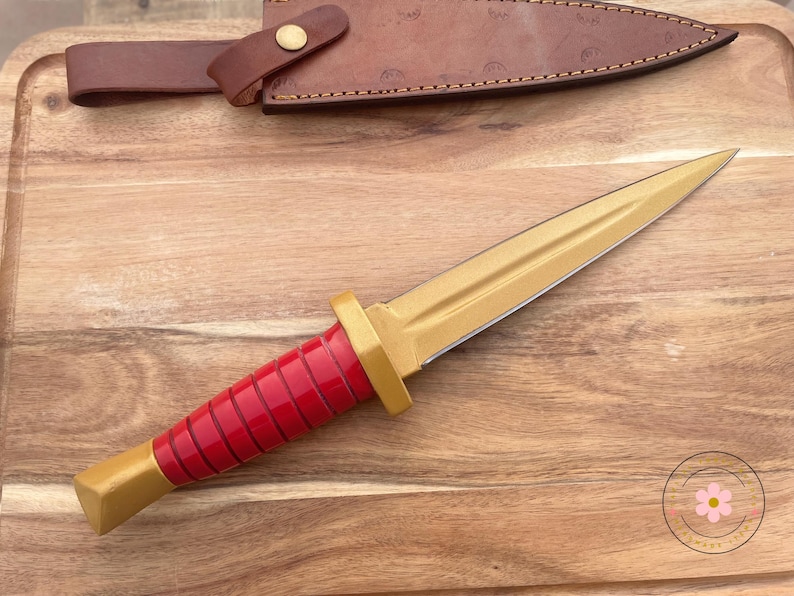 Peut inclure: Un poignard dor&eacute; avec un manche rouge et un &eacute;tui en cuir marron. Le poignard a une lame pointue et un manche d&eacute;coratif. L'&eacute;tui est fix&eacute; avec une sangle en cuir et un bouton en laiton.