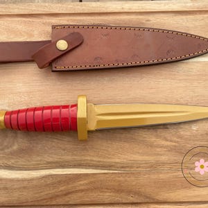 Peut inclure: Un poignard d&eacute;coratif avec une lame dor&eacute;e et un manche rouge. La poign&eacute;e a une base dor&eacute;e et une s&eacute;rie de rainures rouges. Le poignard est pr&eacute;sent&eacute; avec un fourreau en cuir marron et une sangle avec un bouton dor&eacute;. Le poignard et le fourreau sont sur une surface en bois.