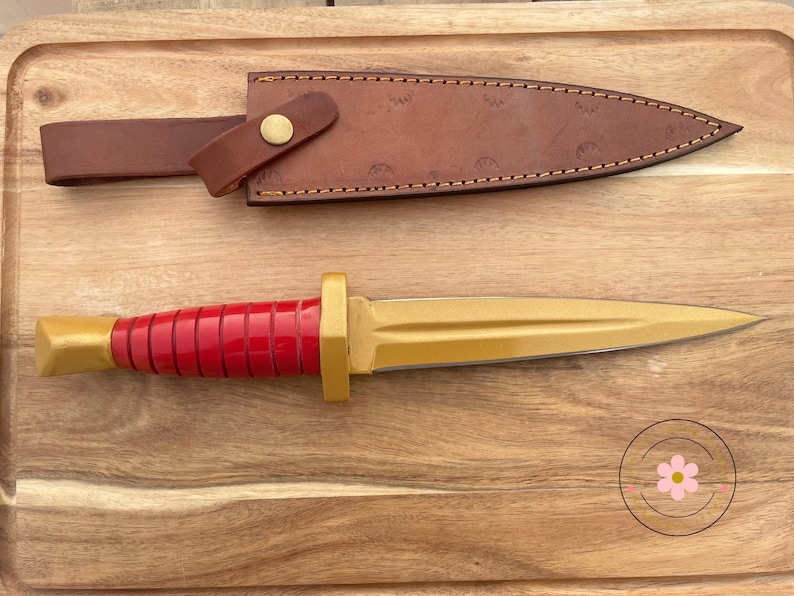Peut inclure: Un poignard d&eacute;coratif avec une lame dor&eacute;e et un manche rouge. La poign&eacute;e a une base dor&eacute;e et une s&eacute;rie de rainures rouges. Le poignard est pr&eacute;sent&eacute; avec un fourreau en cuir marron et une sangle avec un bouton dor&eacute;. Le poignard et le fourreau sont sur une surface en bois.