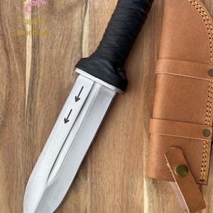 Pu&ograve; includere: Un coltello d'argento con manico in pelle nera e fodero in pelle. Il coltello ha tre frecce che puntano verso il basso sulla lama.