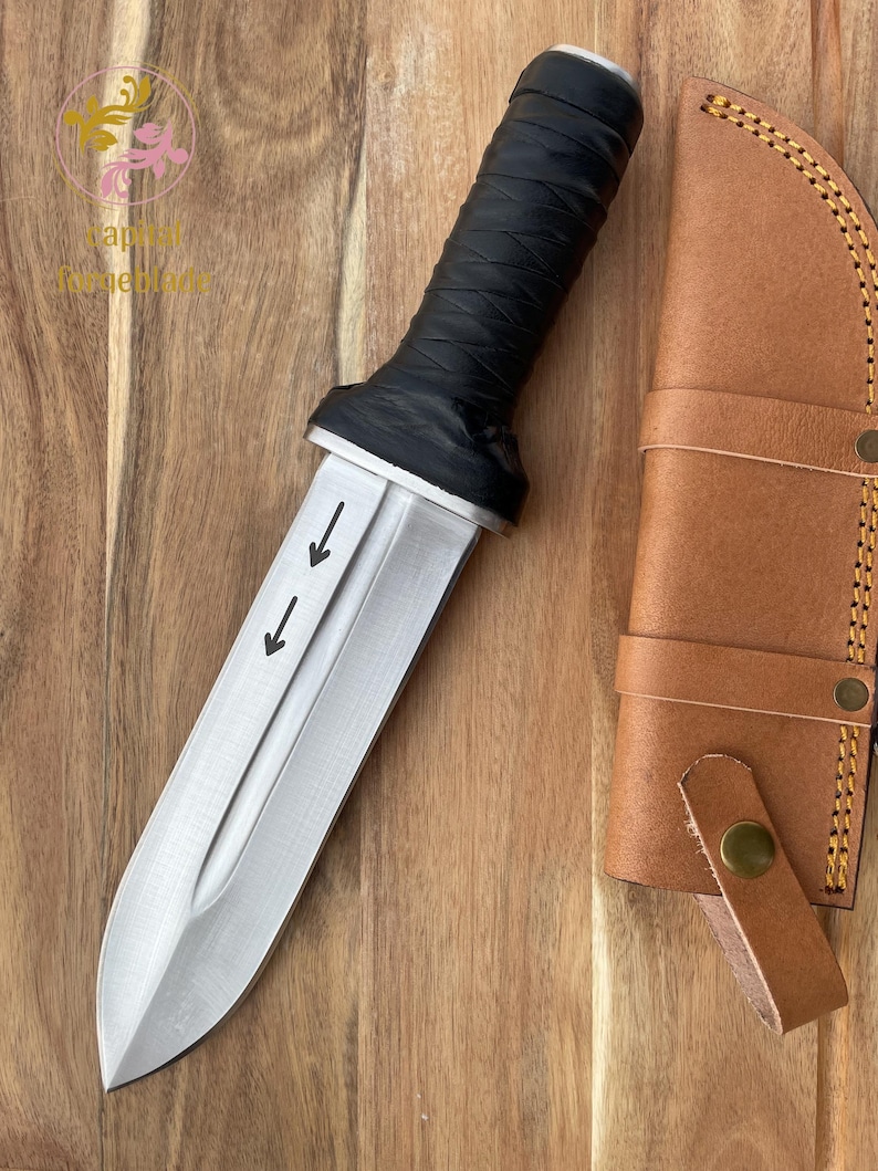 Pu&ograve; includere: Un coltello d'argento con manico in pelle nera e fodero in pelle. Il coltello ha tre frecce che puntano verso il basso sulla lama.