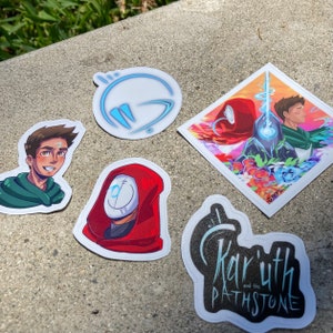 May include: Five stickers featuring illustrations of characters and logos. The stickers are on a gray concrete surface. One sticker features a blue and white logo with the text "Live". Another sticker features a red hooded figure with a white mask. A third sticker features a black and white logo with the text "Kariuth and the Pathstone". A fourth sticker features a colorful illustration of two characters. The fifth sticker features a cartoon character with green hair and a green shirt.