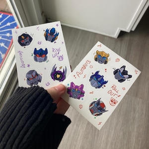 May include: Two sheets of stickers featuring characters from the Transformers franchise. The first sheet features Decepticons with the text "Decepticons" and "Rise Up!" The second sheet features Autobots with the text "Autobots" and "Roll Out!"