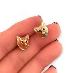 May include: A pair of gold fox head earrings with ruby red eyes. The earrings are shown on a hand.