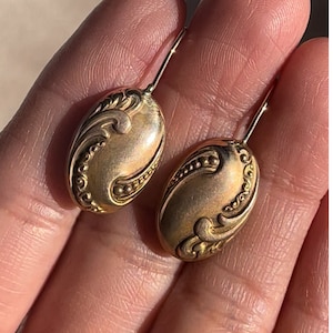 May include: A pair of gold-toned drop earrings with an oval-shaped design. The earrings have a detailed, ornate pattern.