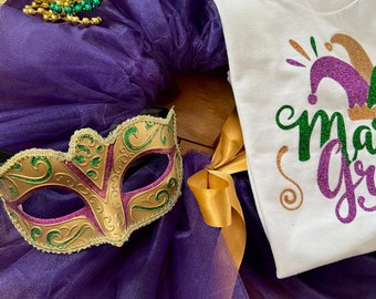 May include: Purple tulle tutu skirt with a gold and green masquerade mask, a gold ribbon, and a white t-shirt with the text "Mardi Gras" in gold glitter.