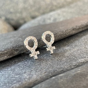 Puede incluir: Un par de pendientes de plata con un diseño de símbolo femenino. Los pendientes están engastados con pequeños cristales transparentes.