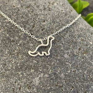 May include: A silver necklace featuring a dainty dinosaur pendant. The dinosaur is outlined in silver and has a simple, modern design.