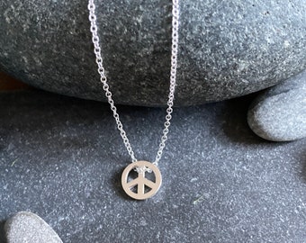 May include: A silver peace sign necklace on a silver chain. The necklace is displayed on a gray stone background.
