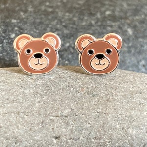 May include: A pair of silver stud earrings featuring a brown bear face design with a pink nose and white eyes.