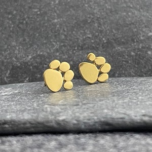 Puede incluir: Un par de pendientes de botón dorados en forma de huellas de patas. Cada huella de pata está formada por varios círculos pequeños.
