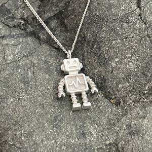 Puede incluir: Collar con colgante de robot de plata. El robot tiene un pecho en forma de corazón con una línea ondulada en el interior. El robot está de pie sobre una superficie de piedra gris.