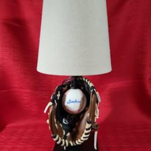 May include: A baseball-themed lamp featuring a baseball glove holding a baseball with the word "Golden" printed on it. The lamp has a beige lampshade and a black base. The background is a solid red color.