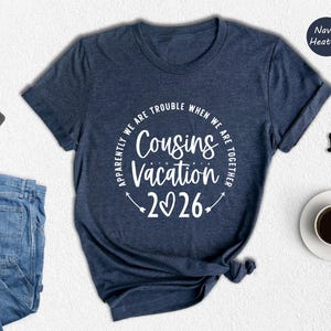a navy blue t-shirt with a humorous text printed on it, along with various items such as a pair of jeans, a wallet, a cup of coffee, and a pair of glasses.