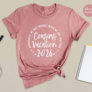 a pink t-shirt with a humorous text printed on it, along with a watch, a notebook, and a pen, all arranged on a beige background.