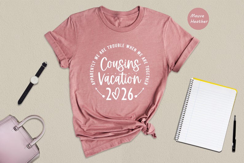 a pink t-shirt with a humorous text printed on it, along with a watch, a notebook, and a pen, all arranged on a beige background.