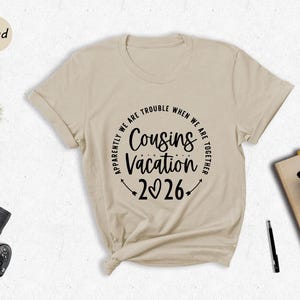 a beige t-shirt with a humorous text design, a potted plant, a camera, a pen, a book, and a cup of coffee, all arranged on a white surface.