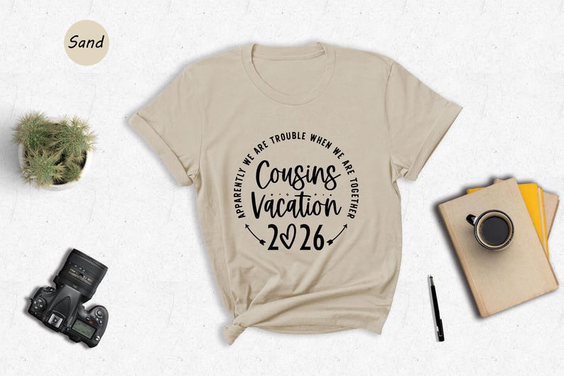 a beige t-shirt with a humorous text design, a potted plant, a camera, a pen, a book, and a cup of coffee, all arranged on a white surface.