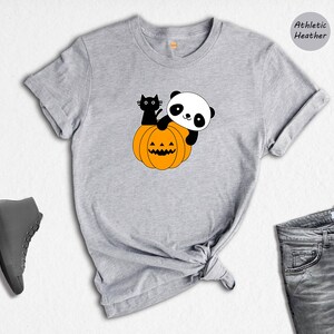 May include: An athletic heather gray t-shirt featuring a graphic of a black cat and a panda sitting on an orange jack-o'-lantern pumpkin. The shirt is displayed alongside a pair of gray sneakers and gray denim jeans.