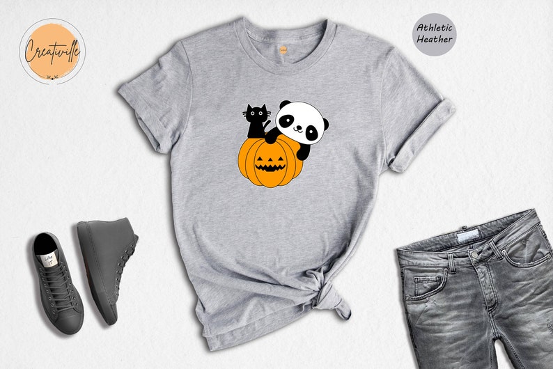 May include: An athletic heather gray t-shirt featuring a graphic of a black cat and a panda sitting on an orange jack-o'-lantern pumpkin. The shirt is displayed alongside a pair of gray sneakers and gray denim jeans.