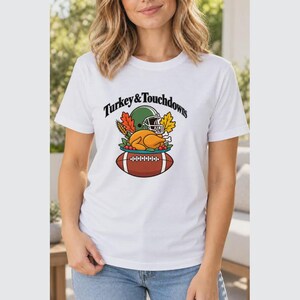 May include: A white short-sleeve t-shirt featuring a graphic of a roasted turkey, a green football helmet, and autumn leaves above a brown football. The text "Turkey & Touchdowns" is printed in black arched lettering above the graphic.