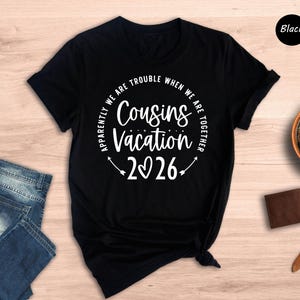 a black t-shirt with a white text design that reads "Apparently we are trouble when we are together" and "Cousins vacation 26". The t-shirt is placed on a wooden surface, along with a pair of blue jeans, a brown wallet, a watch, and a potted plant.