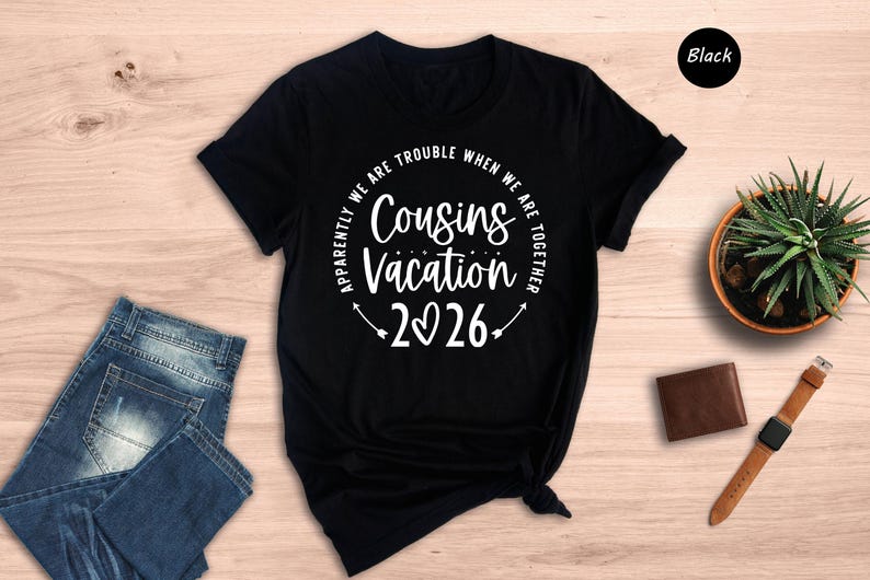 a black t-shirt with a white text design that reads "Apparently we are trouble when we are together" and "Cousins vacation 26". The t-shirt is placed on a wooden surface, along with a pair of blue jeans, a brown wallet, a watch, and a potted plant.