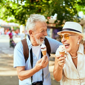 May include: An older couple laughs while enjoying ice cream cones on a sunny day. The man is wearing a light blue shirt and a brown backpack. The woman is wearing a white hat, sunglasses, and a striped shirt.