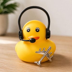 May include: A bright yellow rubber duck wearing a black headset with an attached microphone. The duck features a small orange beak and black eyes. A silver-colored metal airplane figurine is positioned in front of the duck's body.