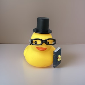 Puede incluir: Un pato de goma amarillo con un sombrero de copa negro y gafas, sosteniendo un pequeño libro azul.