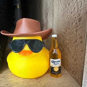 以下が含まれることがあります： 茶色のカウボーイハットとサングラスをかけた黄色のゴム製のダックが、コロナエクストラビールのボトルを持っています。