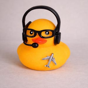 May include: A bright yellow rubber duck featuring a black headset with an attached microphone and black-rimmed glasses. A small, silver-colored airplane emblem is affixed to the front of the duck, which has a distinct orange beak.