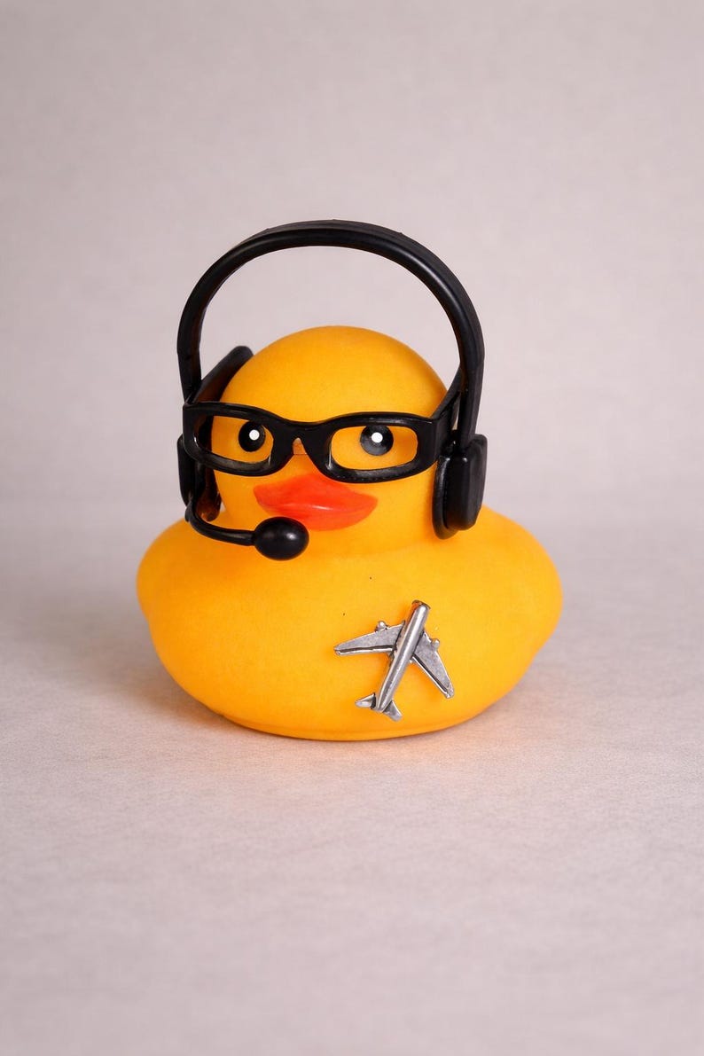 May include: A bright yellow rubber duck featuring a black headset with an attached microphone and black-rimmed glasses. A small, silver-colored airplane emblem is affixed to the front of the duck, which has a distinct orange beak.