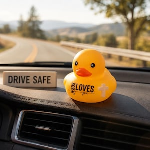 May include: A yellow rubber duck with the text "JESUS LOVES you" and a cross graphic. A small rectangular sign with "DRIVE SAFE" is also visible. The items are on a car dashboard with a scenic road in the background.